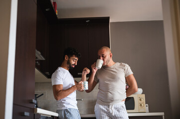 Young interracial gay couple in pajamas having tea in a kitchen. Handsome homosexual men, Caucasian and Mixed race, enjoy morning routine at home. LGBT family everyday lives concept.