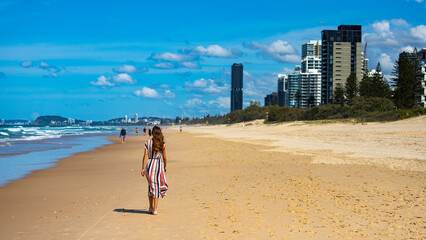 beautiful long-haired girl in long colorful dress walks along beach in gold coast on sunny day; beach with huge skyscrapers; miami beach in gold coast, australia