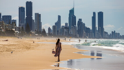 beautiful long-haired girl in long colorful dress walks along beach in gold coast on sunny day; beach with huge skyscrapers; miami beach in gold coast, australia - obrazy, fototapety, plakaty
