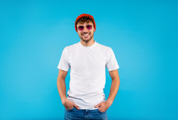 Attractive young smiling man in white t-shirt and orange hat wears glasses stands on blue background