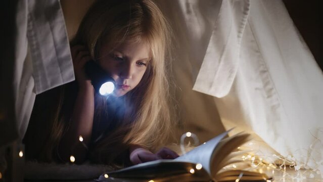 A sweet little girl reads a book with fairy tales at night with a flashlight in the house in the dark sitting in a tent with garlands.