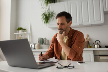 thoughtful man holding hand near face and writing in notebook near laptop in kitchen.