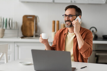 cheerful man in eyeglasses holding coffee cup and talking on mobile phone near blurred laptop.