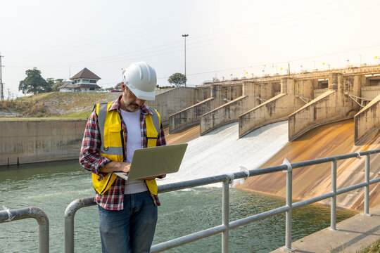 A dam engineering doing his checking routine. He is wearing a white hard hat and yellow transparent vest. He is standing by the rail by the dam.