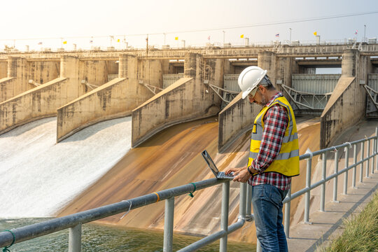 A dam engineering doing his checking routine. He is wearing a white hard hat and yellow transparent vest. He is standing by the rail by the dam.