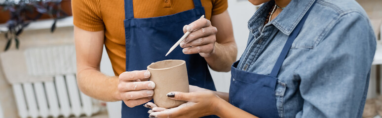 cropped view of african american woman near man in apron modeling clay cup with shaper, banner.