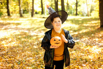 charming boy in a witch costume with a pumpkin in his hands in an autumn park, children for...