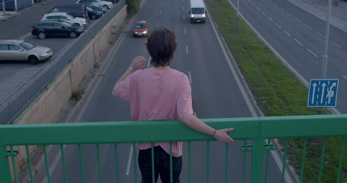 The guy's back, he stands on the bridge. He holds on to the support with his hands. Deciding to jump down. The movement of cars on the road. Dangerous moment.
