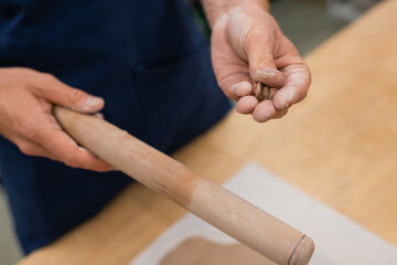 cropped view of man holding wooden rolling pin and piece of clay in hands.