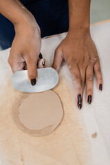 top view of african american woman with manicure holding steel scraper while shaping clay piece.