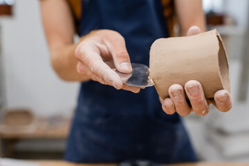 Cropped view of sculptor forming clay product with steel scraper in pottery workshop.