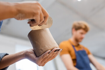 African american woman making clay cup with steel scraper near blurred boyfriend in pottery...