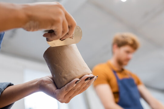 African american woman making clay cup with steel scraper near blurred boyfriend in pottery workshop. - Powered by Adobe