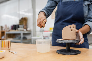 Cropped view of african american sculptor holding sponge with water near clay sculpture in workshop.