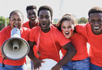African people football supporter fans watching soccer match event at stadium - Multi generational people with red t-shirts having fun on sport world championship event