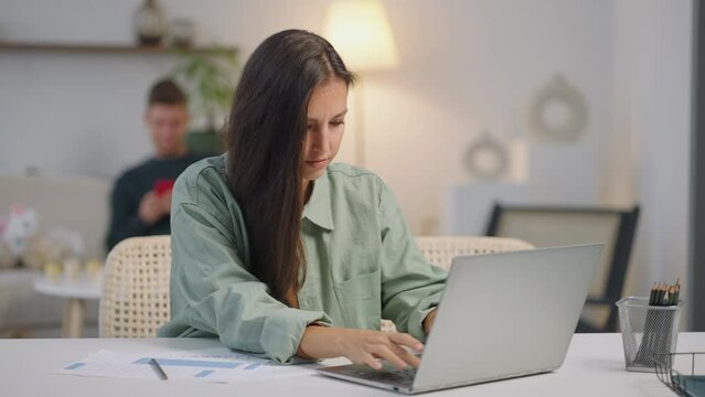 Busy business woman typing on a computer remotely works at the laptop while the father plays and relaxes with the children in the background.