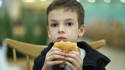 Tired sad boy of seven years old eats a burger at a food court after a school day. Child in sweatshirt enjoying a juicy burger