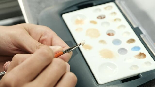 Dental technician coloring dental prosthesis with a paint brush at the laboratory. Aesthetic coloring of teeth. Concept of implantats producing.