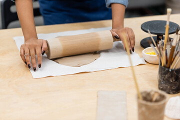partial view of young african american woman in apron modeling clay with rolling pin near paintbrushes.
