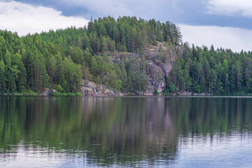 Fototapeta na wymiar Pine forest growing on cliffs by the lake in Repovesi National park on sunny summer day. Beautiful reflection in the calm water 