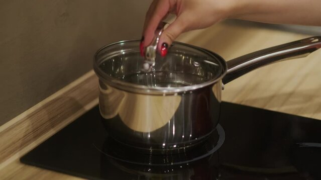 Unrecognizable woman puts pan with water on induction hob, covers with lid in modern kitchen. Concept of cooking.