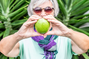Defocused caucasian senior woman showing to the camera a raw green apple standing outdoor in a garden. Concept of healthy eating and stay well