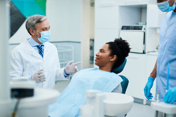 Happy stomatologist talks to black female patient during dental procedure at dentist's office.