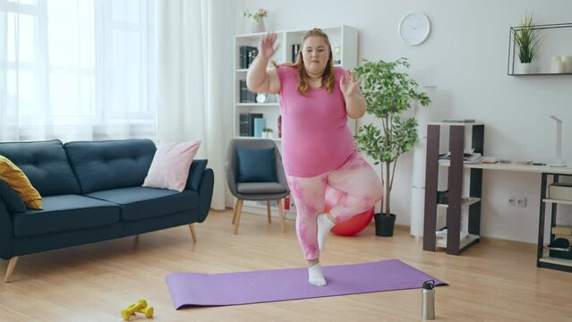 Comic overweight young woman training at home, fails to balance on one leg