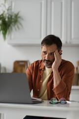 tired man looking at blurred laptop in kitchen and suffering from headache.