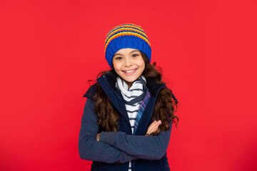 winter fashion. happy kid with curly hair in hat crossed hands. teen girl on red background.