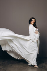 Cute pregnant young slim lady with tummy in spacious room with white walls and large windows.Pregnant girl with curly hair in a long white dress posing for camera.Concept of happy pregnancy.