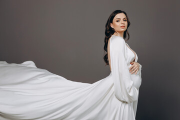 Cute pregnant young slim lady with tummy in spacious room with white walls and large windows.Pregnant girl with curly hair in a long white dress posing for camera.Concept of happy pregnancy.