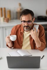 man in eyeglasses holding smartphone and coffee cup while looking at blurred laptop near empty notebook.