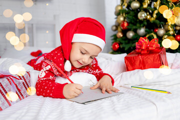 cute beautiful girl child writes a letter to Santa Claus at the Christmas tree in a red sweater and...