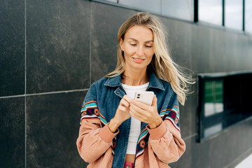 Young stylish woman using phone for online messages while standing outside