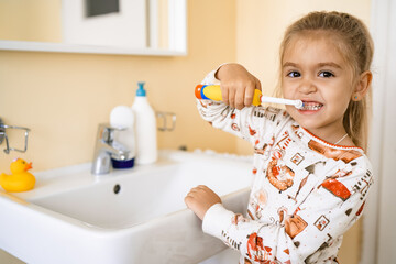 A cute little blonde girl is brushing her teeth with an electric brush, standing in the bathroom near the sink with a mirror.Close-up looks at the camera, copy space - obrazy, fototapety, plakaty