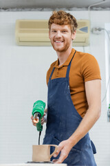 Smiling redhead craftsman in apron holding heat gun near blurred ceramic cup in pottery workshop.