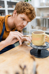 Young redhead craftsman painting on clay cup in pottery workshop.