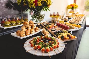 Big table with delicious cocktail of skewers, mini sandwiches, charcuterie, cherry tomato, variety of cheese, olive, salami, artichoke bottoms, nuts, desserts, etc and flowers decoration