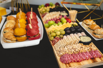 Big wooden board and white plate with salami, crackers, olives, charcuterie, variety of cheeses, variety of mini sandwiches, nuts, etc