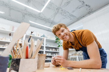 Smiling redhead artisan looking at camera near blurred pottery tools in workshop.