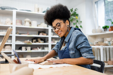 cheerful african american woman in apron and eyeglasses shaping clay piece with hands during pottery class.