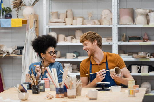 Smiling man looking at african american girlfriend near clay and paintbrushes in pottery studio.