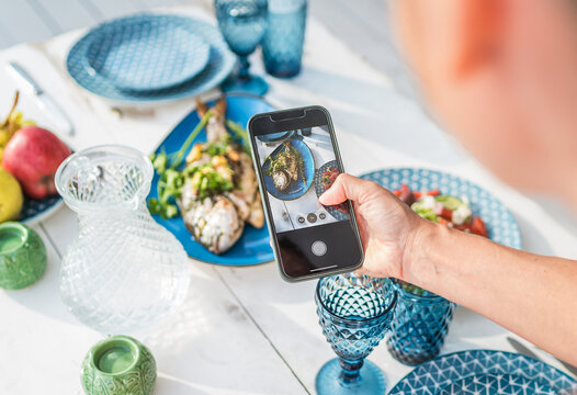 Woman making photo using smartphone Grilled fish in the oven and served on a white wooden table with vegetable salad and fruits. Healthy sea meal preparation, food blogging and serving concept image
