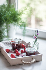 Jars with strawberry jam, strawberries on a linen towel and a white wooden tray and flowers in a vase on the windowsill.