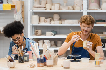 Positive multiethnic couple painting clay sculptures during date in pottery studio.