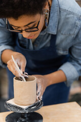 Fototapeta na wymiar African american craftswoman in eyeglasses forming clay cup in workshop.