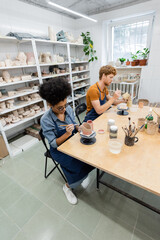 Multiethnic couple in aprons painting on ceramic products in pottery studio.