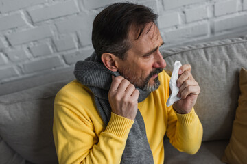 ill man in warm scarf holding paper napkin while sitting on couch at home.