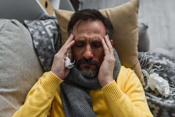 Obraz premium top view of sick man in warm scarf touching head while lying on couch and suffering from migraine.